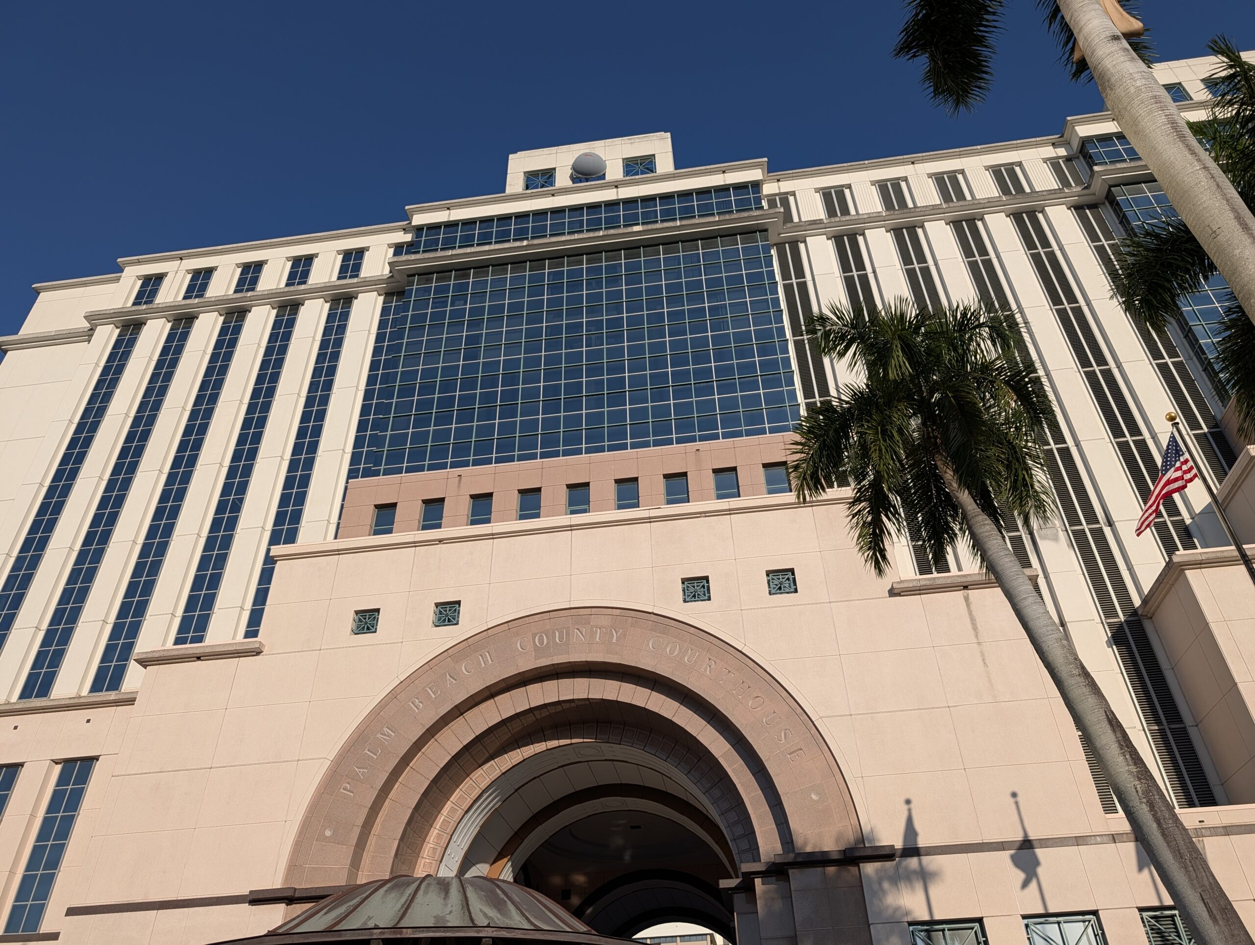 Low-angle exterior view of the Palm Beach County Courthouse in West Palm Beach, showing the arched entrance, glass facade, palm trees and American flag where Palm Beach County probate matters are handled.
