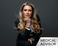 Professional headshot of a woman with long blonde hair in a pinstripe suit, resting her chin on her hand against a dark background